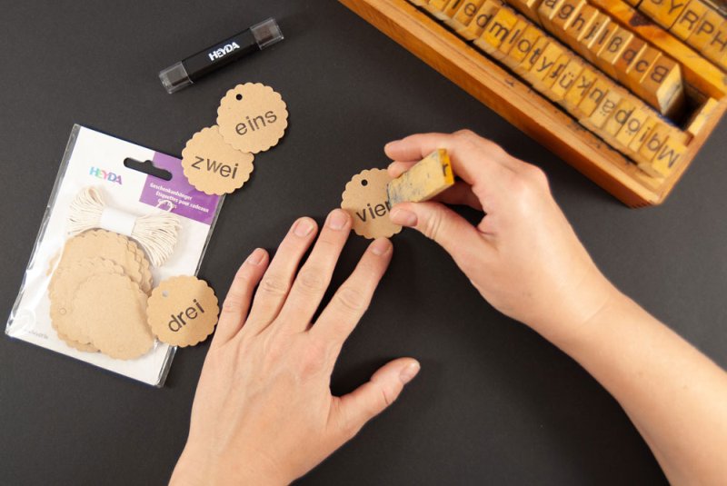 Mit Hilfe eines Stempels werden braune Geschenkanhänger aus Krafpapier der Marke Heyda mit den Worten "eins", "zwei", "drei" und "vier" in schwarzer Tinte bestempelt. Im Hintergrund sieht man einen Kasten, in dem viele verschiedene Buchstaben und Zeichenstempel aufbewahrt sind.