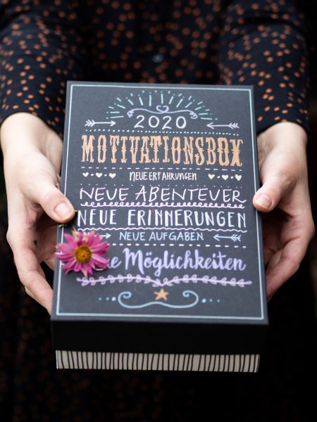 Eine Box wird von einer Person in schwarzen Pulli mit goldenen Punkten gehalten. Die Box wurde mit Metallic Markern von Stabilo belettert. Darauf ist zu lesen "2020 Motivationsbox Neue Erfahrungen, Neue Abenteuer, Neue Erinnerungen, Neue Aufgaben, Neue Möglichkeiten". Auf der Box liegt eine rosafarbene Blume.