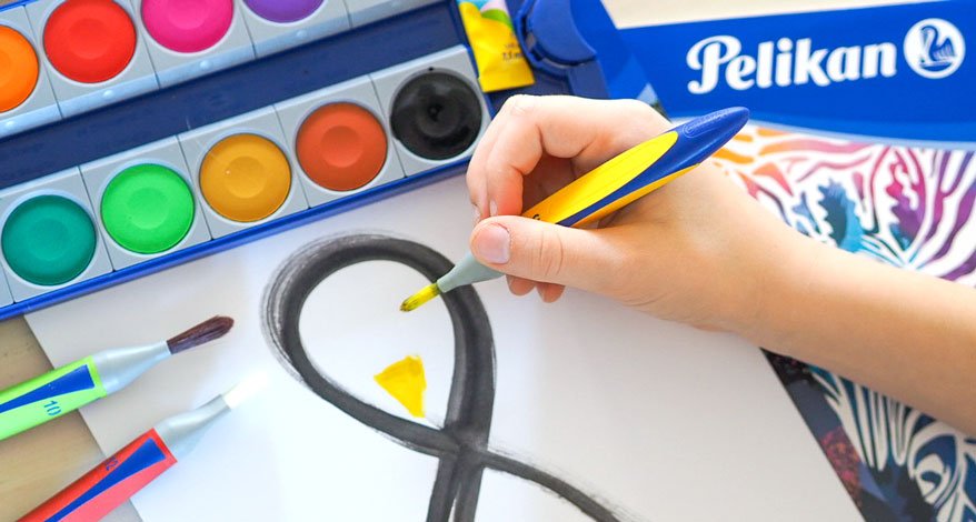 Kinderhand malt mit Wasserfarben auf ein Papier, daneben ein Farbkasten, Pinsel und Zeichenblock von Pelikan