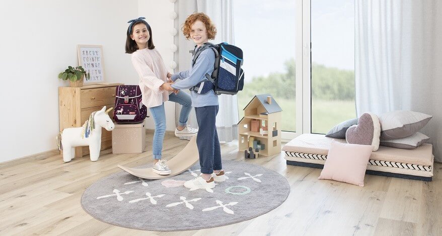 Ein Junge und ein Mädchen stehen in einem Kinderzimmer und lächeln in die Kamera. Der Junge trägt einen blauen Step by Step Schulranzen auf dem Rücken, ein lila Step by Step Schulranzen steht hinter dem Mädchen auf einem Hocker. Es stehen eine Kommode, ein kleines Puppenhaus, ein Plüsch-Einhorn und eine kleine Matratze mit mehreren Kissen im Raum vor dem bodentiefen Fenster.