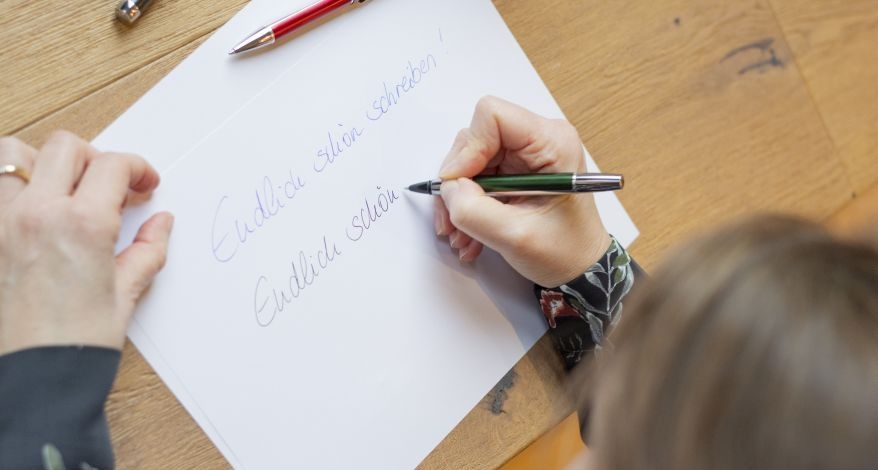 Hand, die mit Rollerball Pura von Pelikan "Endlich schön schreiben!" auf ein Blatt Papier schreibt.