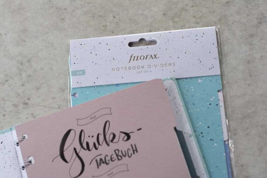 Ein Notizbuch von Filofax liegt aufgeklappt auf einem Schreibtisch, zu sehen ist ein selbstgemachter Divider, auf dem "Glückstagebuch" im Handletteringstil geschrieben steht. Dahinter liegt eine Packung gekaufter Divider von Filofax.
