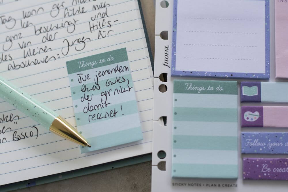 Nahaufnahme eines Notizbuchs von Filofax. Zu sehen ist ein Tagebucheintrag, ein Kugelschreiber und ein eingeklebter Haftzettel auf dem geschrieben steht "Tue jemandem etwas Gutes, der gar nicht damit rechnet!"