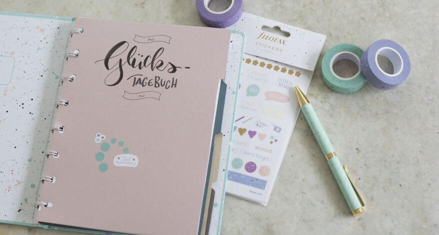 Ein Notizbuch von Filofax liegt aufgeklappt auf einem Schreibtisch, zu sehen ist ein selbstgemachter Divider, auf dem "Glückstagebuch" steht. Daneben liegt ein Stickerbogen, ein Kugelshreiber und Washitape.
