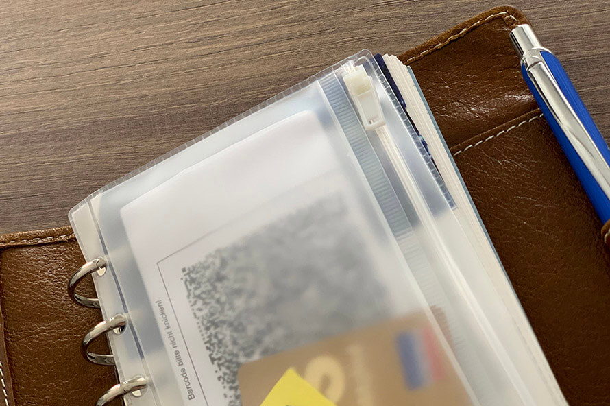 In einem aufgeklappten Kalender der Marke Filofax ist eine befüllte Klarsichttasche mit Reißverschluss und ein blauer Kugelschreiber in der Stiftschlaufe zu sehen.