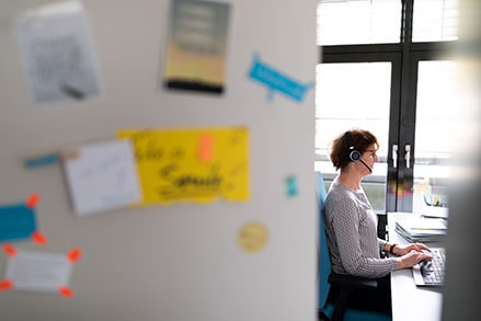 Man sieht durch eine geöffnete Tür in ein Büro. Dort sitzt eine Frau vor einer Tastatur. Sie trägt ein Headset und telefoniert.