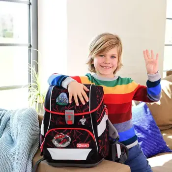 Ein Junge in einem regenbogenfarbenen Pullover sitzt im Wohnzimmer auf dem Sofa und winkt in die Kamera. Stolz drückt er mit der anderen Hand seinen neuen Schulranzen der Marke Step-by-Step an sich.