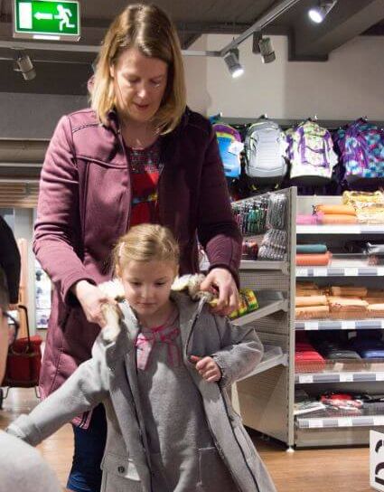 Während einer Schulranzen Beratung zieht ein Mädchen die dicke Winterjacke über, um damit den Sitz des ergobag zu testen. Ein Mitarbeiter hockt vor dem Kind, die Mutter steht dahinter und hilft ihr in die Jacke.