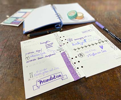 Im Vordergrund liegt ein ausfgefülltes und beklebtes Kalenderblatt mit eingetragenen Terminen. Im Hintergrund liegen Haftklebezettel und ein Notizbuch der Marke Filofax