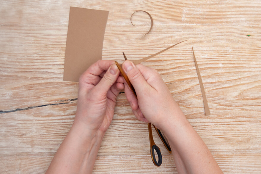 Auf einer hellen Holzoberfläche zieht jemand einen spitzen Streifen braunen Fotokartons von Artoz über eine goldene Schere, um ihn zu rund zu formen. Foto: Johanna Rundel