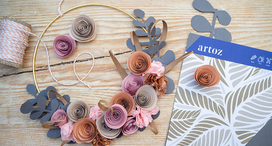 Auf einer hellen Holzoberfläche liegt ein goldener Kranz mit verschiedenen Papierblumen. Daneben liegen eine Packung Papiere von Artoz sowie eine Spule rosa-weißes Bäckergarn. Foto: Johanna Rundel