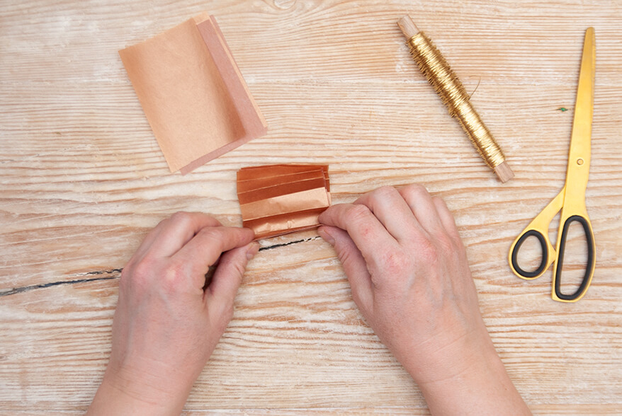 Auf einer hellen Holzoberfläche faltet jemand mehrere Schichten braunes Seidenpapier von Artoz zu einer Ziehharmonika. Daneben liegt eine Spule goldener Draht und eine goldene Schere. Foto: Johanna Rundel