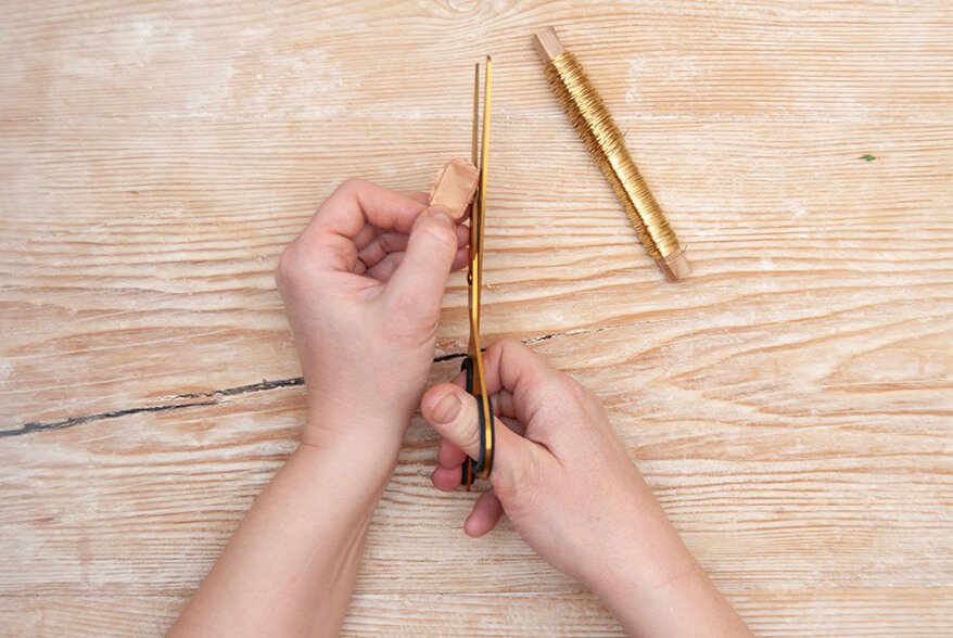 Auf einer hellen Holzoberfläche schneidet jemand einen zur Ziehharmonika gefaltetes Seidenpapier von Artoz an den Enden rund ab. Daneben liegt eine Spule goldener Draht. Foto: Johanna Rundel