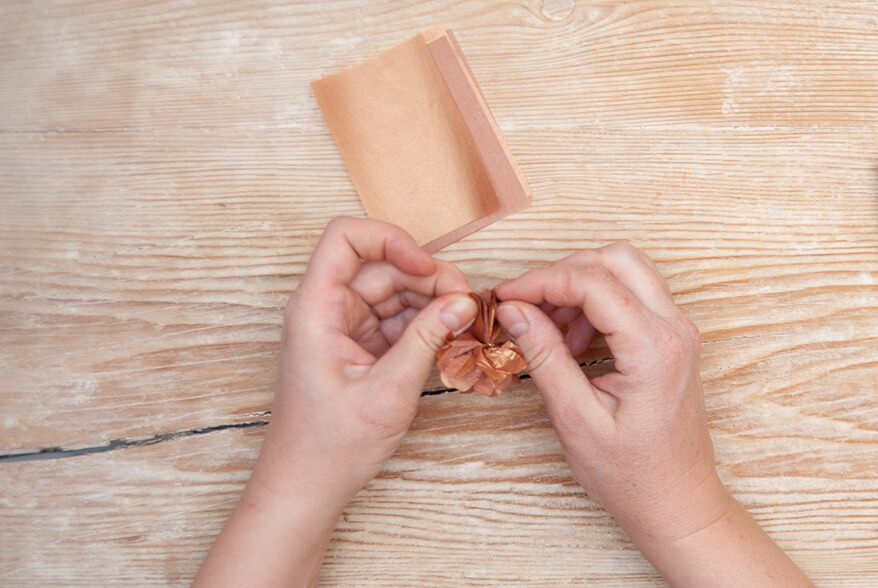Auf einer hellen Holzoberfläche fächert jemand einen zur Ziehharmonika gefaltetes Seidenpapier von Artoz zu einem Pompon auf. Foto: Johanna Rundel