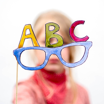 Ein Mädchen im Grundschulalter, mit blonden Haaren und roten Kleidern, hält ein buntes Foto-Prop in Form einer Brille, mit den Buchstaben ABC darauf, in die Kamera. Der verwendete Fotokarton ist von der Marke Heyda. Foto: Johanna Rundel
