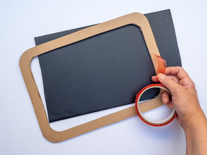 Jemand klebt einen Rahmen aus brauner Pappe auf ein, mit Tafelfolie von Knorr-Prandell bezogenes Stück Pappe, um daraus eine kleine Schultafel als Foto-Prop für die Einschulung zu basteln. Foto: Johanna Rundel