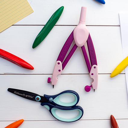 Auf einem weißen Untergrund liegen eine blaue Schere und ein lila Zirkel aus der griffix-Serie von Pelikan. Drumherum sind bunte Wachsmalkreiden verteilt. Foto Johanna Rundel