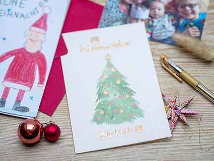 Auf einem Holztisch liegt eine Weihnachtskarte von ABC Grußkarten, auf der in gold "Frohe Weihnachten und ein gutes neues Jahr" geschrieben steht. Außerdem ist ein handgemalter, geschmückter Weihnachtsbaum zu sehen. Drumherum liegen ein selbstgemaltes Weihnachtsbild, ein Familienfoto, ein gefalteter Papierstern, Weihnachtsbaumkugeln und ein Goldstift. Foto: Johanna Rundel