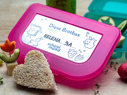 Auf einem Holzbrett liegen eine pinke und eine türkise Schulbrotdose von School-Mood. Auf den Aufkleber der pinken Dose wurden Name und Klasse eines Mädchens geschrieben. Vor der Brotdose liegt ein in Herzform ausgestochenes Toastsandwich und ein Käsespieß. Foto: Johanna Rundel