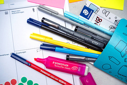 Auf einem hellgrauen Untergrund liegen verschiedene Materialien, die man zur Durchführung eines Workshops benötigt: unterschiedliche Stifte und Marker von Staedtler in einer blauen Box, bunte Klebepunkte und Haftnotizzettel sowie ein ausgedruckter Workshop-Canvas zur Vorbereitung. Foto: Johanna Rundel