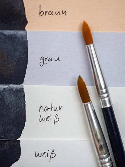 Drei verschiedenfarbige Aquarellpapiere von Hahnemühle liegen übereinander. Rechts daneben stehen die Farbtöne: braun, grau, naturweiß und weiß. Foto: Johanna Rundel 