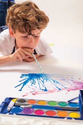 Ein Junge liegt auf dem Boden und hat einen Trinkhalm im Mund. Er pustet damit flüssige Wassermalfarbe über ein Papier, die zu einem kleinen Kunstwerk zerfließt. Im Vordergrund liegt ein Deckfarbkasten von Pelikan.