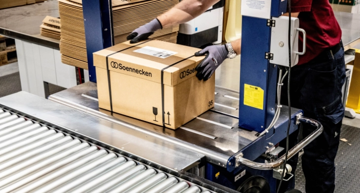 Arbeiter mit Schutzhandschuhen sichert Karton mit Soennecken Aufdruck auf einer Förderanlage in einer Verpackungsstation.