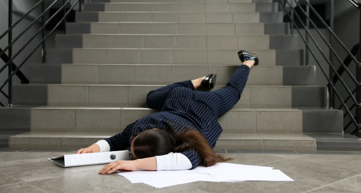 Frau in Businesskleidung nach Treppensturz, verstreute Dokumente auf Boden.