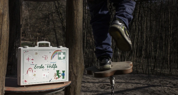 Erste-Hilfe-Koffer für Kinder auf Spielplatz, Kind balanciert auf Holzplattform.