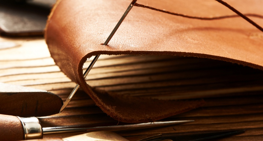 Ein Stück Leder wird verarbeitet, es steckt eine Nadel in einem Stück Leder. Daneben liegt ein Werkzeug zur Bearbeitung.