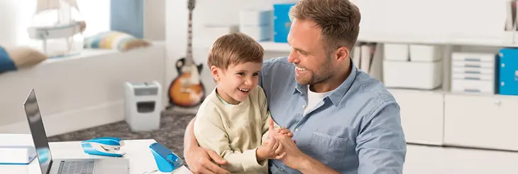Ein Vater mit Kind, auf einem Schreibtisch ein Laptop, ein Locher und Tacker von Leitz.