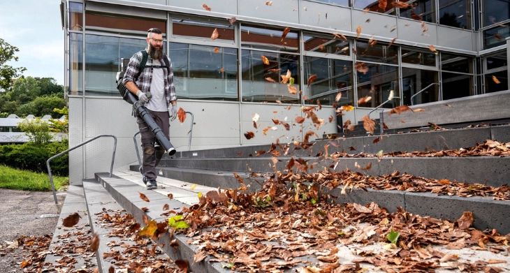 Mann entfernt mit einem Kärcher Laubbläser Herbstlaub von einer Treppe vor einem modernen Gebäude.