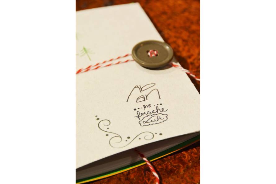 Auf den Umschlag eines Notizbuchs aus Karton sind Elemente und Text gelettert. Das Notizbuch ist mit einem Knopf und Garn verschlossen.