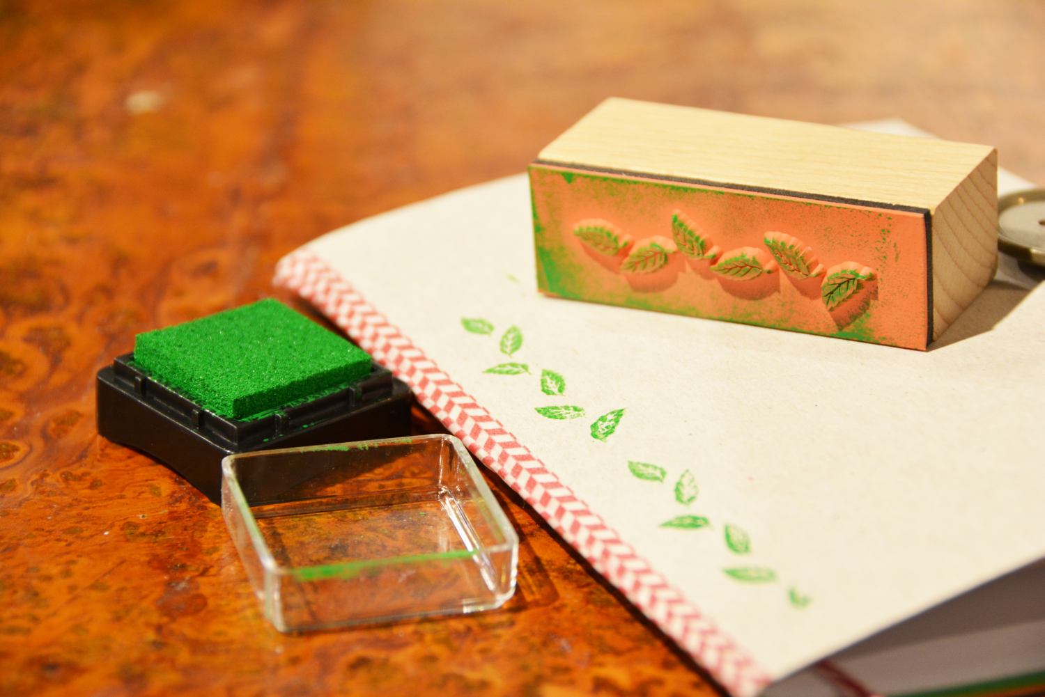 Auf einem Notzibuch aus Karton liegt ein Stempel mit Blättermotiv. Dieses ist mit grüber Stempelfarbe auf das Notizbuch gestempelt. Das Stempelkissen liegt offen daneben.