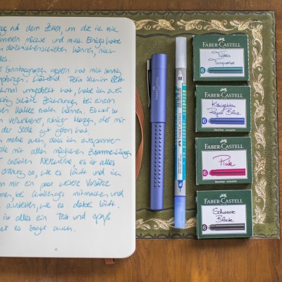 Neben einem aufgeschlagenen und beschriebenen Tagebuch liegt ein Grip-Füller der Marke Faber-Castell, ebenso wie ein Tintenlöscher und mehrere unterschiedlich farbige Tintenpartonen-Verpackungen von Faber-Castell.