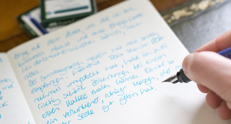 Ein aufgeschlagenes Tagebuch, in das jemand gerade etwas mit Füller reinschreibt. Im Hintergrund sind Tintenpatronen Verpackungen von Faber-Castell zu sehen.