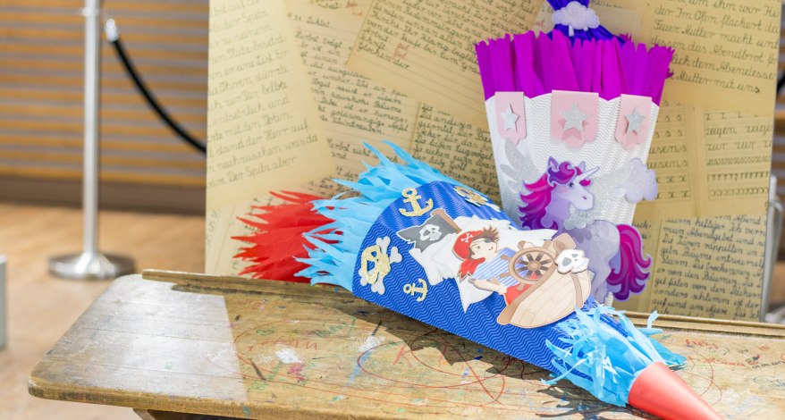 2 bunte Schultüten der Marke Heyda, eine in blau mit Piraten-Motiv, die andere in lila mit Einhorn, liegen auf einer alten Schulbank.