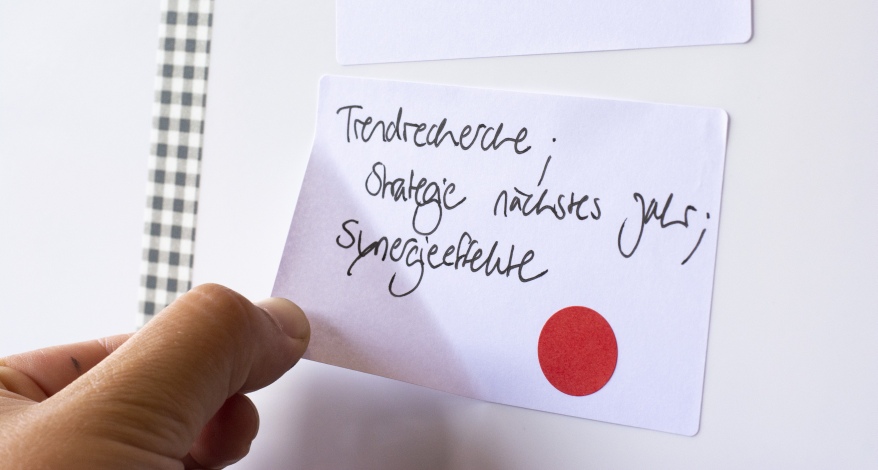 Eine Hand zieht ein Etikett mit der Aufschrift "Trendrecherche; Strategie nächstes Jahr, Synergieeffekte" von einem Flip Chart. Auf dem Zettel klebt ein roter Punkt.