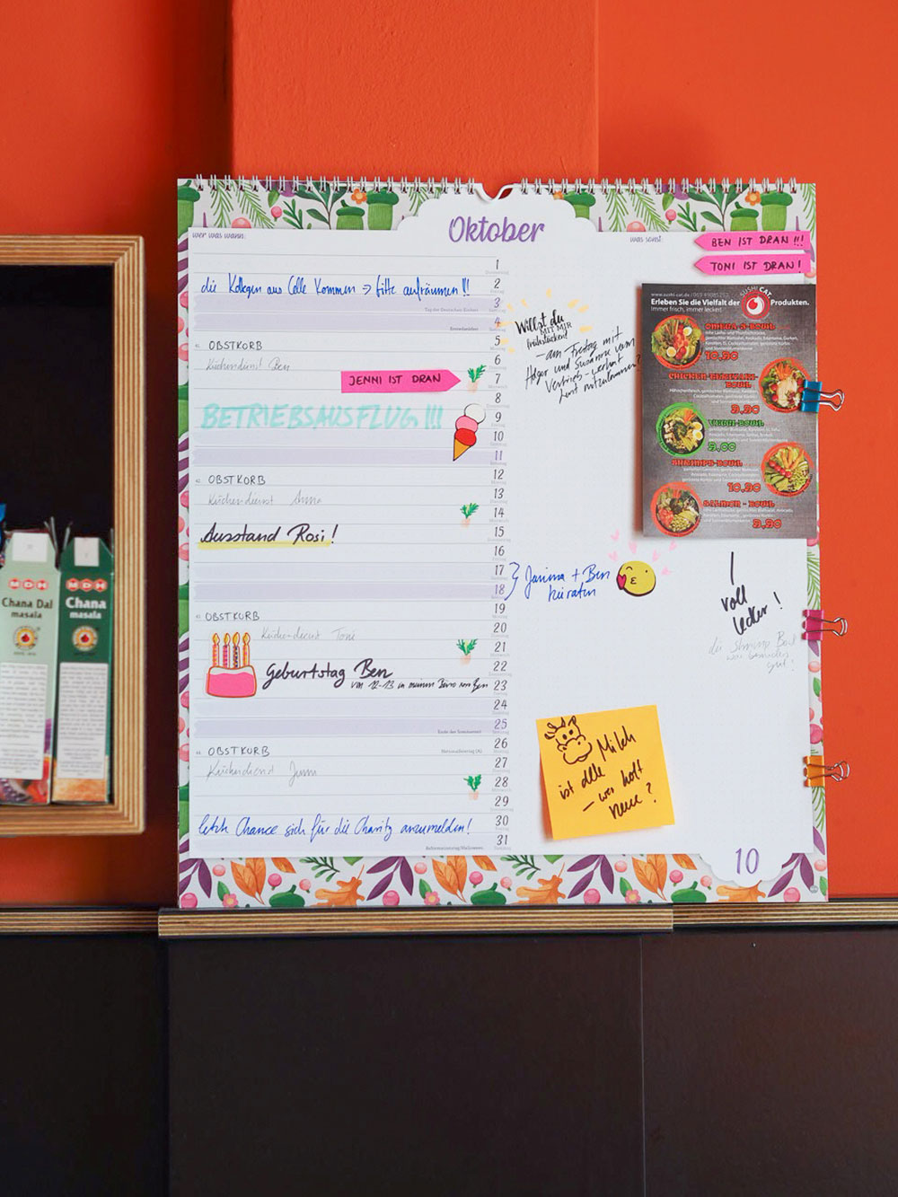Ein großer bunter Planner mit florelem Motiv der Marke Athesia ist zu sehen. Er steht vor einer roten Wand. Der Planer ist mit einigen Flyern und Haftnotizen beklebt und wurde mit einigen Terminen beschriftet. 