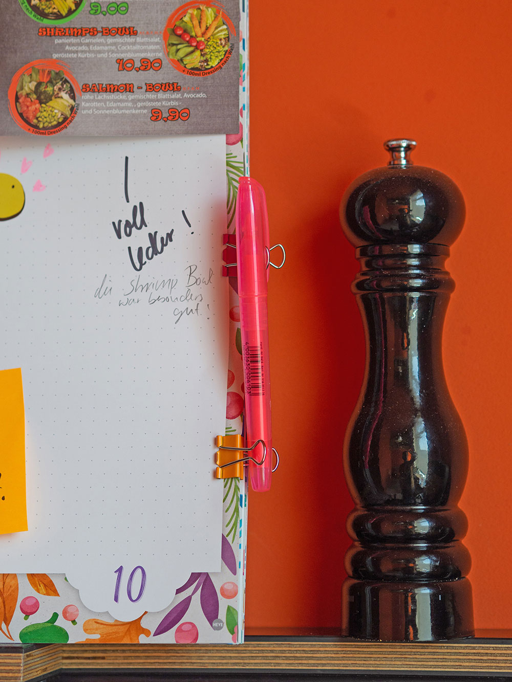An einen Planner der Marke Athesia wurde mit Hilfe von zwei Foldback-Klammern ein rosa Textmarker befestigt. Rechts neben dem Kalender steht eine große Pfeffermühle. Der Hintergrund ist orange. 