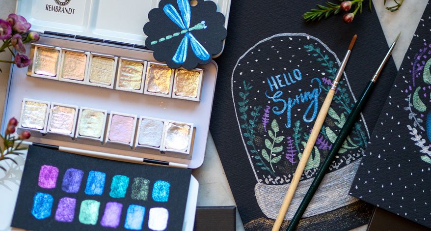 Schimmernde Aquarellfarben mit Pinseln und Frühlingsmotiv auf schwarzem Papier.