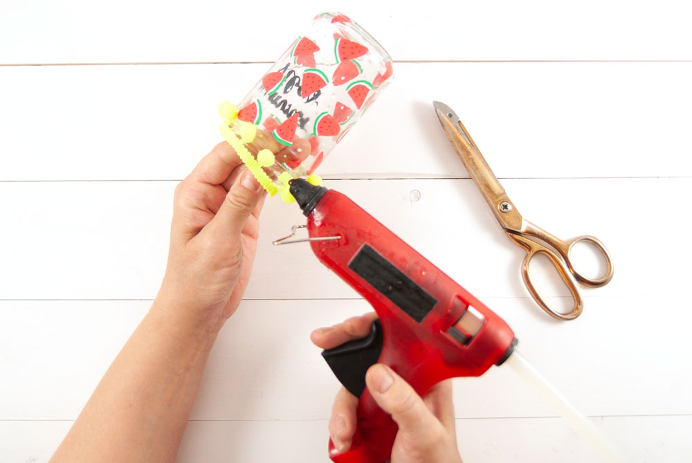 Mit einer Heißklebepistole wir ein Bommelband an ein Glas mit Melonenmuster geklebt