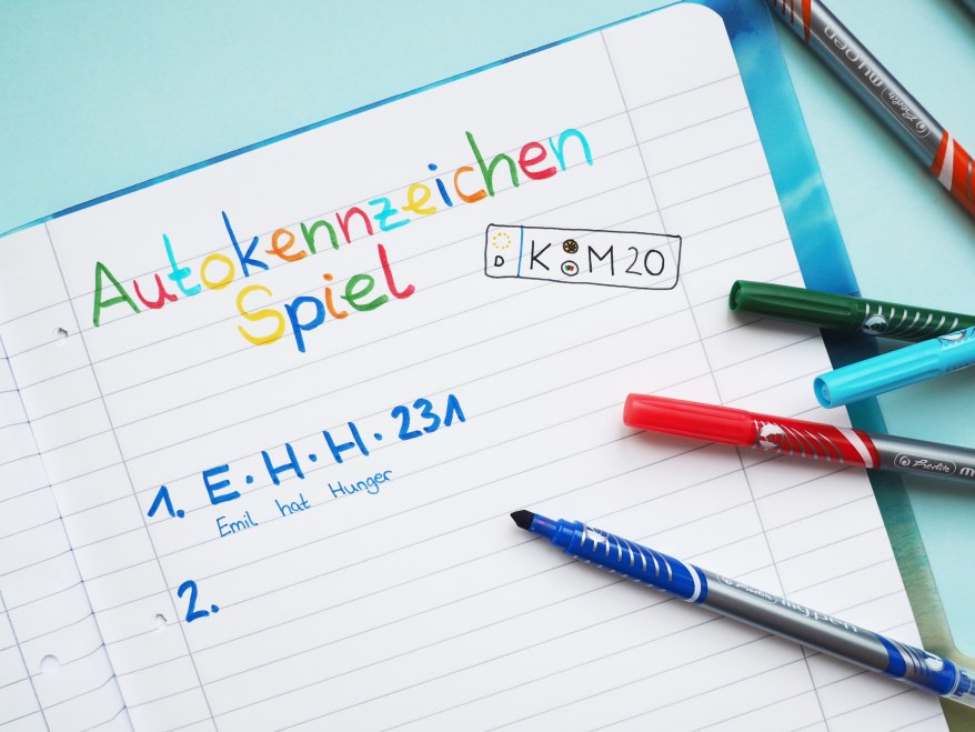 Schreibheft mit bunter Überschrift "Autokennzeichen-Spiel" und bunten Stiften daneben.