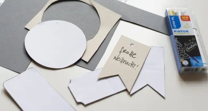 Auf einem Schreibtisch entstehen Geschenkanhänger zu Weihnachten mit Schablonen und aus Papierresten.