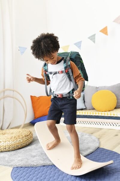 Ein Junge steht im Kinderzimmer auf einer Wippe und balanciert darauf. Auf dem Rücken trägt er einen Step by Step Schulranzen.