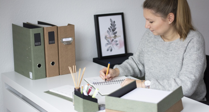 Frau arbeitet im Homeoffice und sitzt am Schreibtisch. Sie schreibt in ein Notizbuch und ist umgeben von Büromaterialien aus der S.O.H.O.-Serie von Rössler.
