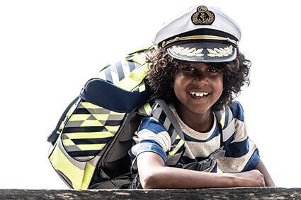 Ein Junge mit Lockenkopf und Kapitänsmütze steht hinter einer Holzmauer und stützt sich mit einem Arm auf. Er trägt einen Schulranzen der Firma School-Mood.