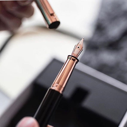 Im Zentrum des Bildes ist ein schwarzer Füller mit roségoldenen Akzenten der Marke Otto Hutt, der von einer Hand gehalten wird. In der andern Hand ist die Kappe des Füllers zu sehen. 