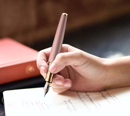 Im Anschnitt ist eine Hand zu sehen, die mit einem Roségoldfarbenenen Füller der Marke Otto Hutt in eine Notizbuch schreibt. Im Hintergrund liegen zwei Bücher. 