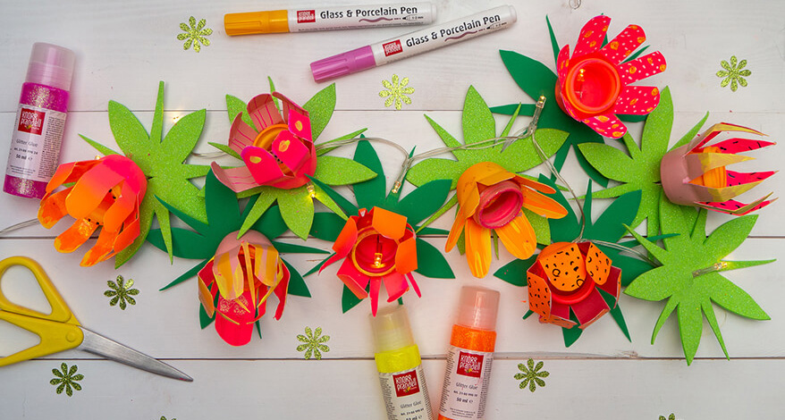 Auf einem hellen Untergrund liegt eine Lichterkette aus upcycelten Plastikflaschen in Blumenform. Die Farben der Blüten sind Orange, Pink, Rosa und Gelb. Unter den Blüten sind grüne Blätter aus Moosgummi. Die Glühbirnchen der Lichterkette leuchten. Drumherum liegen verschiedene Glass & Porcelain Pens und Glitzerkleber von Knorr Prandell und eine gelbe Schere. Foto: Johanna Rundel